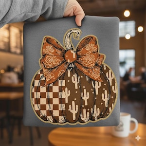 May include: A gray pillow featuring a brown pumpkin design with a decorative bow. The pumpkin has a checkered pattern and cactus illustrations. The bow has a floral design. The pillow is held by a person.