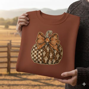 May include: A rust-colored sweatshirt featuring a pumpkin design. The pumpkin has a checkered pattern, cactus illustrations, and a bow. The bow and pumpkin stem are in shades of brown and orange.