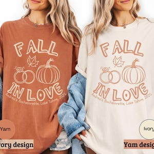 May include: Two t-shirts, one yam-colored and one ivory, feature the text "FALL IN LOVE" with a leaf, rings, and pumpkin graphic. The text "Andrea's Bachelorette, Lake Tahoe, 2025" is also printed on the shirts. The shirts are part of a fall-themed collection.