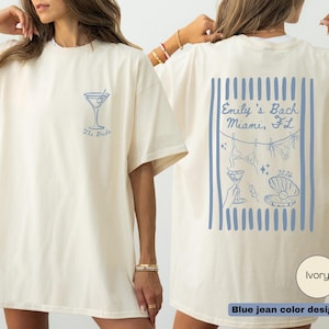 May include: Ivory-colored t-shirt with a blue cocktail glass graphic and the text "The Bride" on the front. The back features a blue design with the text "Emily's Bach Miami, FL" and other illustrations. The shirt is a blue jean color design.