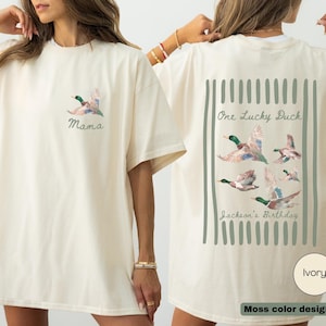 May include: Ivory-colored t-shirt with a duck graphic. The front features a flying duck and the word "Mama." The back displays a design with multiple ducks, the text "One Lucky Duck" and "Jackson's Birthday."