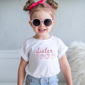 Op de afbeelding: Een jong kind draagt een wit t-shirt met het woord "Sister" in roze letters en aardbeienillustraties. Het kind draagt ook een roze zonnebril en een roze haarband. Het kind lacht.