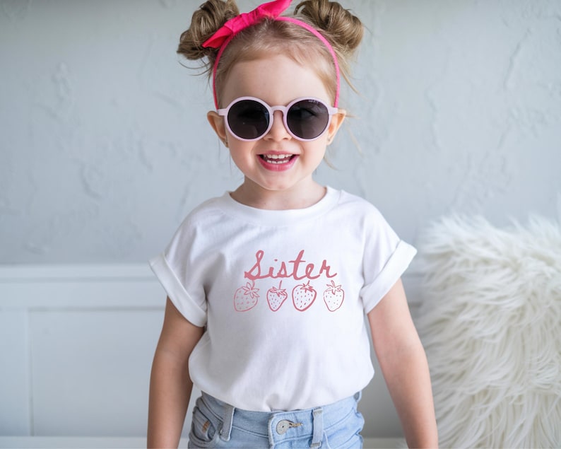 Op de afbeelding: Een jong kind draagt een wit t-shirt met het woord "Sister" in roze letters en aardbeienillustraties. Het kind draagt ook een roze zonnebril en een roze haarband. Het kind lacht.