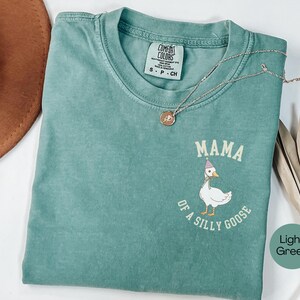 Może przedstawiać: Jasnozielona koszulka Comfort Colors z napisem "MAMA of a silly goose" i kresk&oacute;wkową gęsią w czapce imprezowej. Złoty naszyjnik z wisiorkiem znajduje się na koszulce.