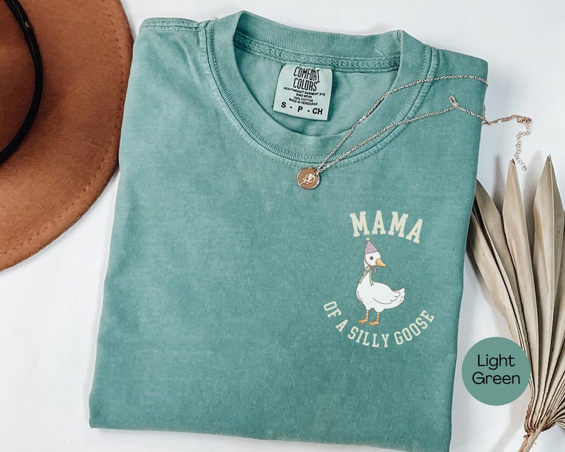 Może przedstawiać: Jasnozielona koszulka Comfort Colors z napisem "MAMA of a silly goose" i kresk&oacute;wkową gęsią w czapce imprezowej. Złoty naszyjnik z wisiorkiem znajduje się na koszulce.