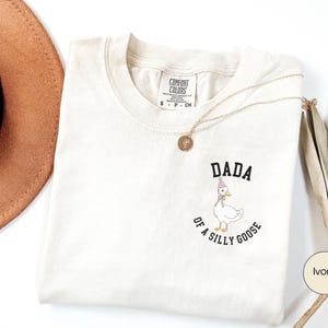 Może przedstawiać: Kremowa koszulka z napisem "DADA of a SILLY GOOSE" i kresk&oacute;wkową gęsią w czapeczce. Koszulka jest złożona i ma metkę Comfort Colors. Widoczny jest r&oacute;wnież złoty naszyjnik i brązowy kapelusz.