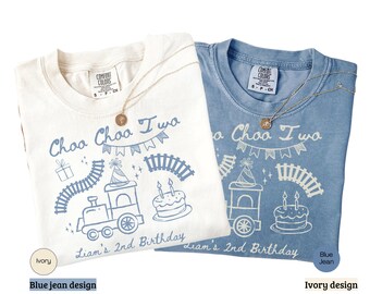 Könnte beinhalten: Zwei T-Shirts, eines in Elfenbein und eines in Blau, mit einem Geburtstagsthema zum Thema Zug. Die Shirts zeigen den Text "Choo Choo Two" und "Liam's 2nd Birthday" mit Illustrationen von Zug, Kuchen und Geschenk. Jedes Shirt hat eine goldene Halskette.