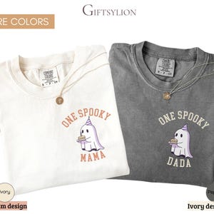May include: Two short-sleeve crew neck t-shirts in ivory and pepper gray. Each shirt features a graphic of a ghost wearing a party hat and holding a cake. The ivory shirt has the text ONE SPOOKY MAMA, and the pepper gray shirt has the text ONE SPOOKY DADA.