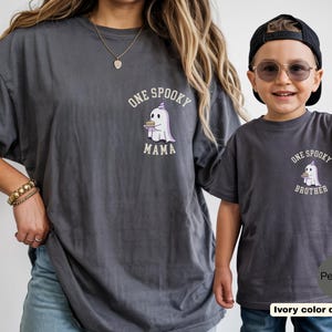 May include: Two charcoal gray t-shirts featuring a graphic of a ghost wearing a party hat and holding a small cake. The larger shirt has the text "ONE SPOOKY MAMA" and the smaller shirt has the text "ONE SPOOKY BROTHER" printed in white lettering.
