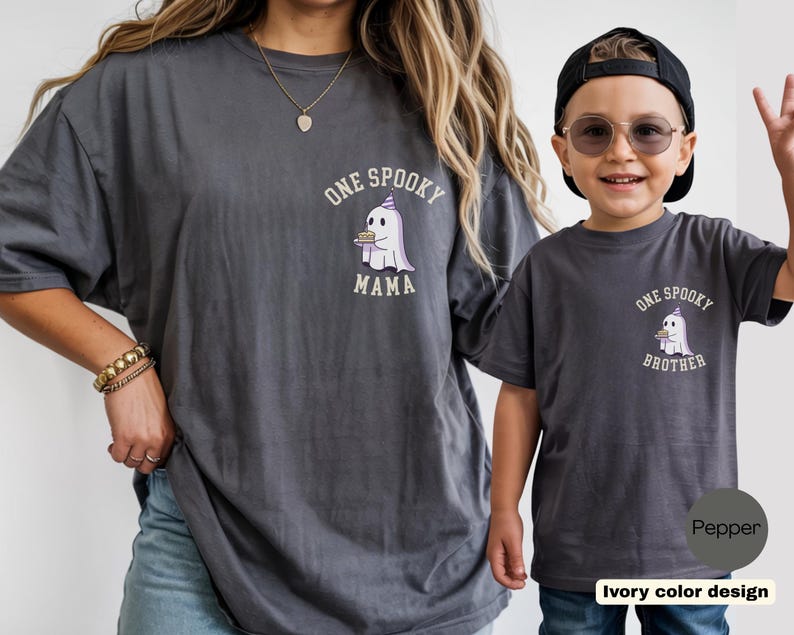 May include: Two charcoal gray t-shirts featuring a graphic of a ghost wearing a party hat and holding a small cake. The larger shirt has the text "ONE SPOOKY MAMA" and the smaller shirt has the text "ONE SPOOKY BROTHER" printed in white lettering.