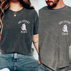 May include: A charcoal gray crew neck t-shirt featuring a small graphic on the left chest of a white ghost wearing a purple party hat and holding a cake. The text above the ghost reads ONE SPOOKY and below it reads MAMA or DADA in white.