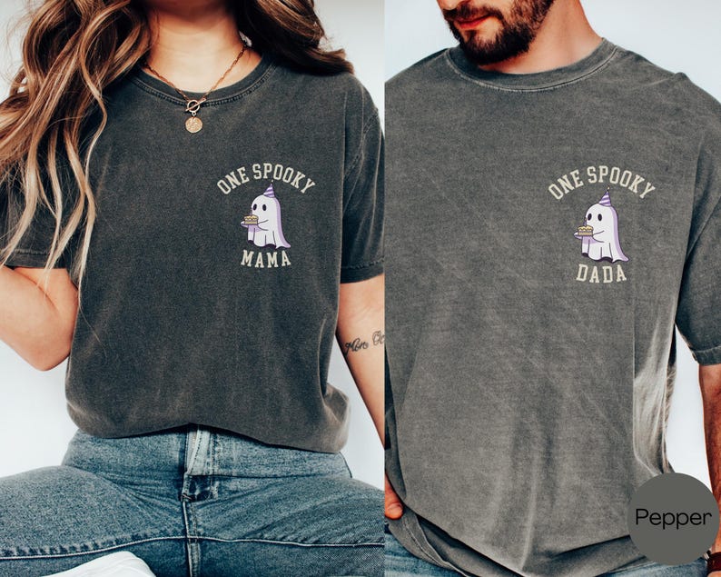 May include: A charcoal gray crew neck t-shirt featuring a small graphic on the left chest of a white ghost wearing a purple party hat and holding a cake. The text above the ghost reads ONE SPOOKY and below it reads MAMA or DADA in white.