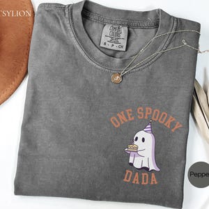 May include: A charcoal gray short-sleeve t-shirt with a Comfort Colors label at the neck. The chest features a graphic of a white ghost wearing a purple party hat and holding a birthday cake, with the orange text ONE SPOOKY DADA surrounding it.