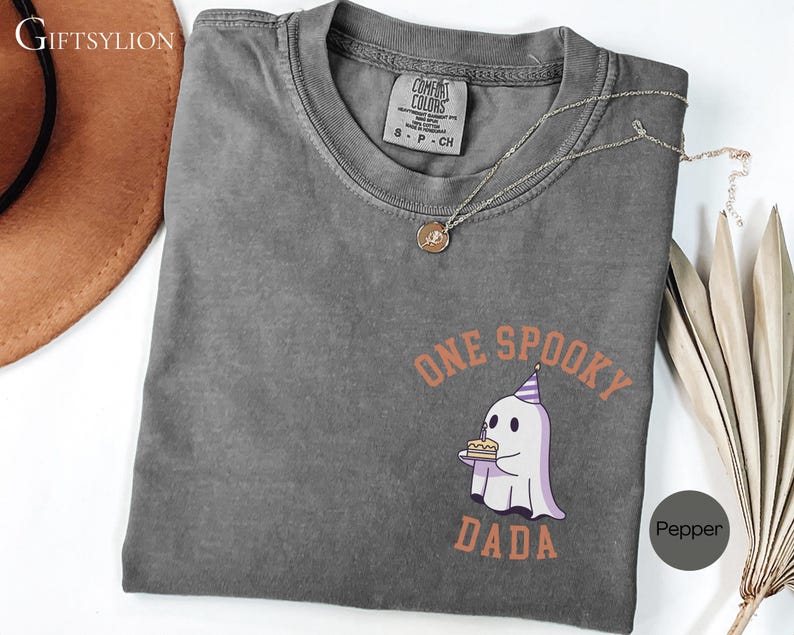 May include: A charcoal gray short-sleeve t-shirt with a Comfort Colors label at the neck. The chest features a graphic of a white ghost wearing a purple party hat and holding a birthday cake, with the orange text ONE SPOOKY DADA surrounding it.