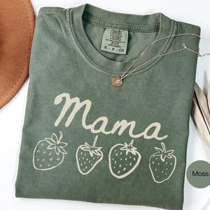 Op de afbeelding: Een mosgroen t-shirt met het woord "Mama" in cursief en vier aardbeienillustraties. Het shirt heeft een ronde hals en een klein label. Een gouden ketting met een hanger is over het shirt gedrapeerd.