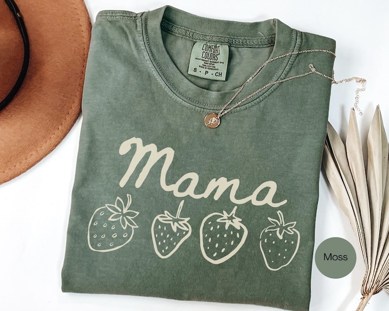 Op de afbeelding: Een mosgroen t-shirt met het woord "Mama" in cursief en vier aardbeienillustraties. Het shirt heeft een ronde hals en een klein label. Een gouden ketting met een hanger is over het shirt gedrapeerd.