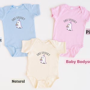 May include: Three baby bodysuits in light blue, pink, and natural colors. Each garment features a graphic of a white ghost wearing a purple party hat and holding a small cake, with the text ONE SPOOKY arched above the ghost in dark gray letters.