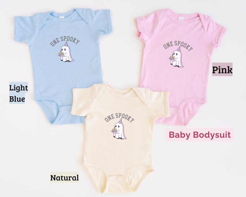 May include: Three baby bodysuits in light blue, pink, and natural colors. Each garment features a graphic of a white ghost wearing a purple party hat and holding a small cake, with the text ONE SPOOKY arched above the ghost in dark gray letters.