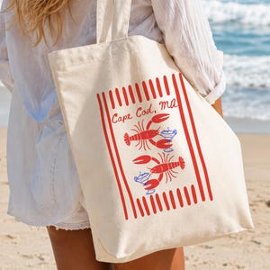 May include: A cream-colored canvas tote bag featuring a red vertical striped border and the text Cape Cod, MA. The design includes two red lobster illustrations, each positioned next to a blue and white cocktail glass with a lime garnish.