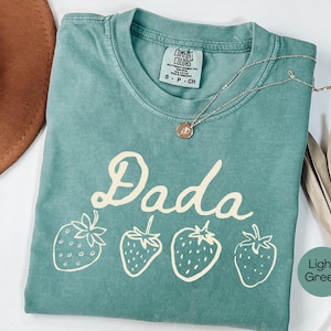 Op de afbeelding: Een lichtgroen t-shirt met het woord "Dada" in een speels lettertype en vier aardbeienillustraties. Het shirt heeft een ronde hals en korte mouwen. Een gouden ketting met een hanger is over het shirt gedrapeerd.