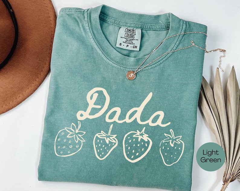 Op de afbeelding: Een lichtgroen t-shirt met het woord "Dada" in een speels lettertype en vier aardbeienillustraties. Het shirt heeft een ronde hals en korte mouwen. Een gouden ketting met een hanger is over het shirt gedrapeerd.