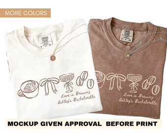 May include: Two Comfort Colors t-shirts, one cream and one brown, with a graphic design. The design includes a coffee cup, bow, champagne glass, and coffee beans. Text reads "Love is Brewing Ashley's Bachelorette". Each shirt has a gold pendant necklace.