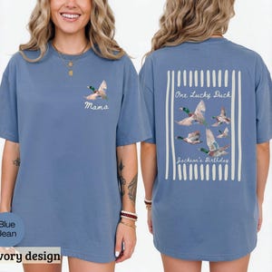May include: Blue jean colored t-shirt with a duck graphic. The front of the shirt has the word "Mama" and a duck graphic. The back of the shirt has the text "One Lucky Duck" and "Jackson's Birthday" with a duck graphic.