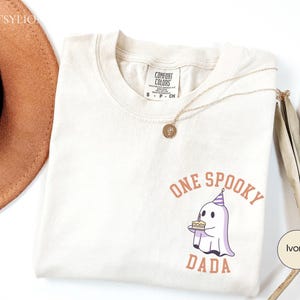 May include: An ivory short-sleeve t-shirt featuring a graphic of a ghost wearing a party hat and holding a birthday cake. The text above and below the ghost reads ONE SPOOKY DADA in a curved orange font. The shirt has a Comfort Colors brand label.
