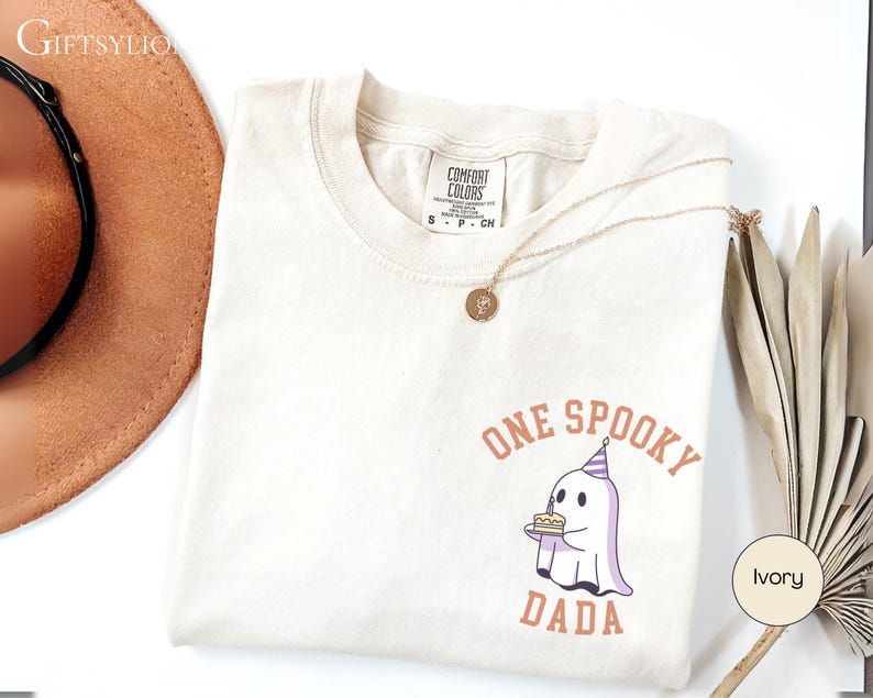 May include: An ivory short-sleeve t-shirt featuring a graphic of a ghost wearing a party hat and holding a birthday cake. The text above and below the ghost reads ONE SPOOKY DADA in a curved orange font. The shirt has a Comfort Colors brand label.