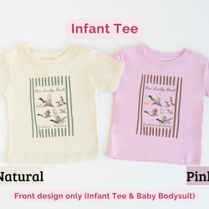 May include: Two infant tees, one natural and one pink, are displayed. Each tee features a graphic of flying ducks with the text "One Lucky Duck" and "Jackson's Birthday" within a vertical striped frame. The text "Infant Tee" is above.
