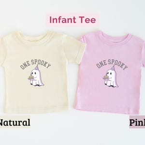 May include: Two short-sleeve infant tees in natural and pink colors. Each shirt features a graphic of a white ghost wearing a purple party hat and holding a birthday cake, with the text "ONE SPOOKY" arched above the ghost in gray lettering.