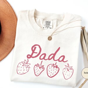 Op de afbeelding: Ivoor kleurig T-shirt met het woord "Dada" in een cursief lettertype en vier roze aardbeien illustraties. Het shirt heeft een ronde hals en een "Comfort Colors" label. Een gouden ketting en een bruine hoed zijn ook zichtbaar.