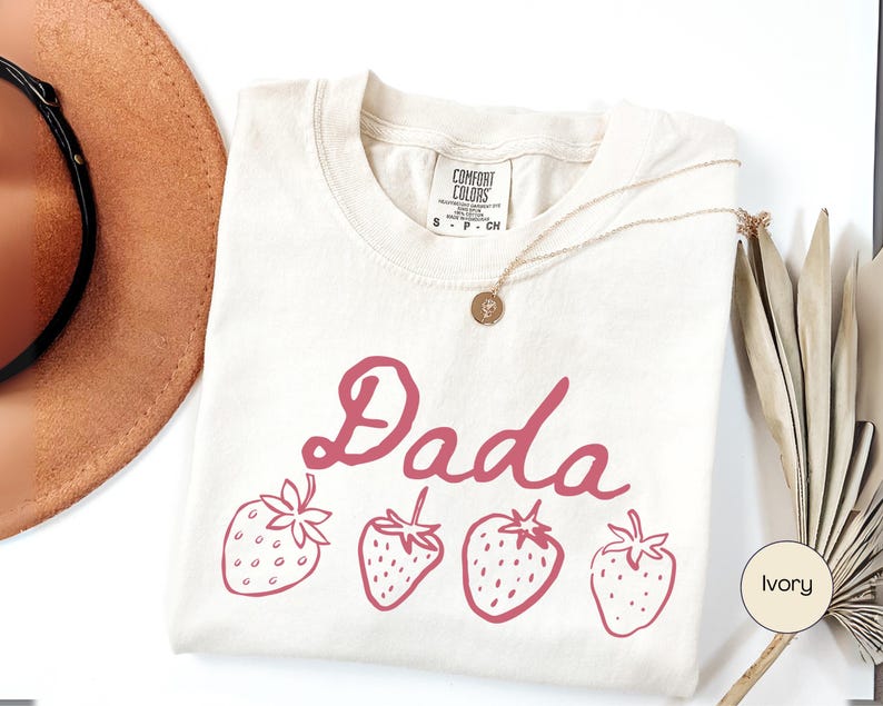 Op de afbeelding: Ivoor kleurig T-shirt met het woord "Dada" in een cursief lettertype en vier roze aardbeien illustraties. Het shirt heeft een ronde hals en een "Comfort Colors" label. Een gouden ketting en een bruine hoed zijn ook zichtbaar.