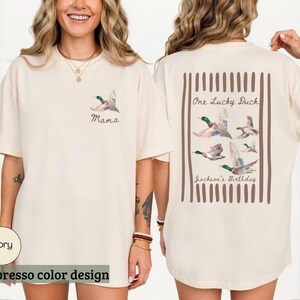 May include: Ivory-colored t-shirt with an espresso color design. The front features a watercolor duck and the word "Mama." The back displays a rectangular design with ducks and the text "One Lucky Duck" and "Jackson's Birthday."