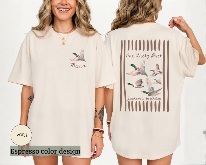 May include: Ivory-colored t-shirt with an espresso color design. The front features a watercolor duck and the word "Mama." The back displays a rectangular design with ducks and the text "One Lucky Duck" and "Jackson's Birthday."