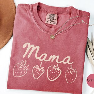 Op de afbeelding: Karmizijnrood T-shirt met het woord "Mama" in cursief en vier aardbeienillustraties. Een zilveren ketting met een hanger ligt op het shirt. Het shirt heeft een Comfort Colors label.