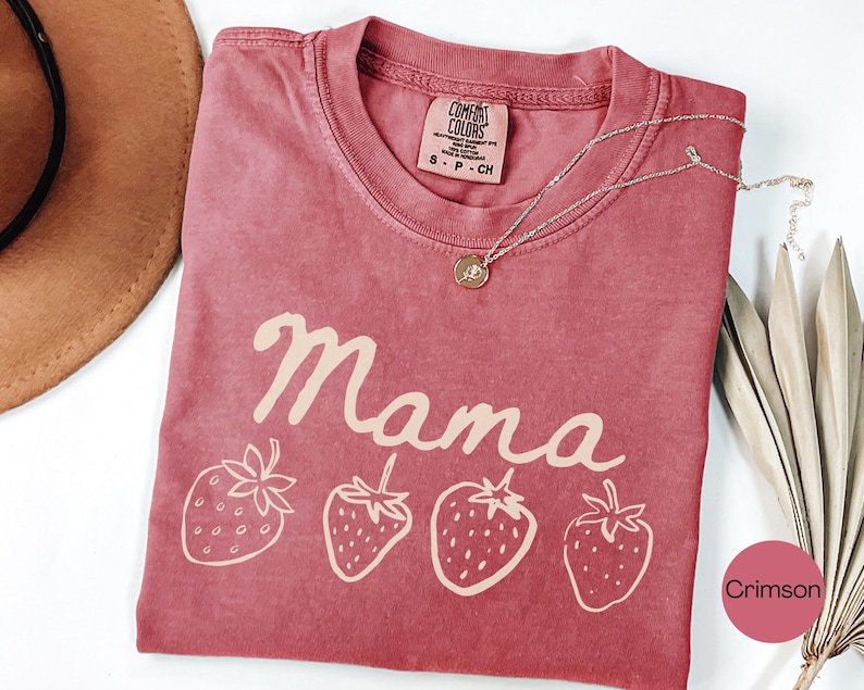Op de afbeelding: Karmizijnrood T-shirt met het woord "Mama" in cursief en vier aardbeienillustraties. Een zilveren ketting met een hanger ligt op het shirt. Het shirt heeft een Comfort Colors label.