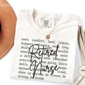 May include: Ivory-colored t-shirt with the words "Retired Nurse" in a bold, black script. The shirt features a list of words related to nursing, such as "care," "comfort," and "heal." A gold necklace with a pendant rests on the shirt.