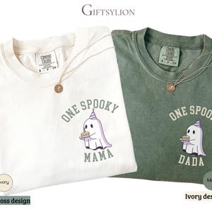 May include: An ivory and a moss green t-shirt, each featuring a graphic of a ghost wearing a purple striped party hat and holding a birthday cake. The ivory shirt has the text ONE SPOOKY MAMA, and the moss green shirt has the text ONE SPOOKY DADA.