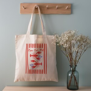 May include: A cream-colored canvas tote bag featuring a red vertical striped rectangular border. Inside the border are two red lobster illustrations and the text Cape Cod, MA in red script. The bag has two long shoulder straps for carrying.