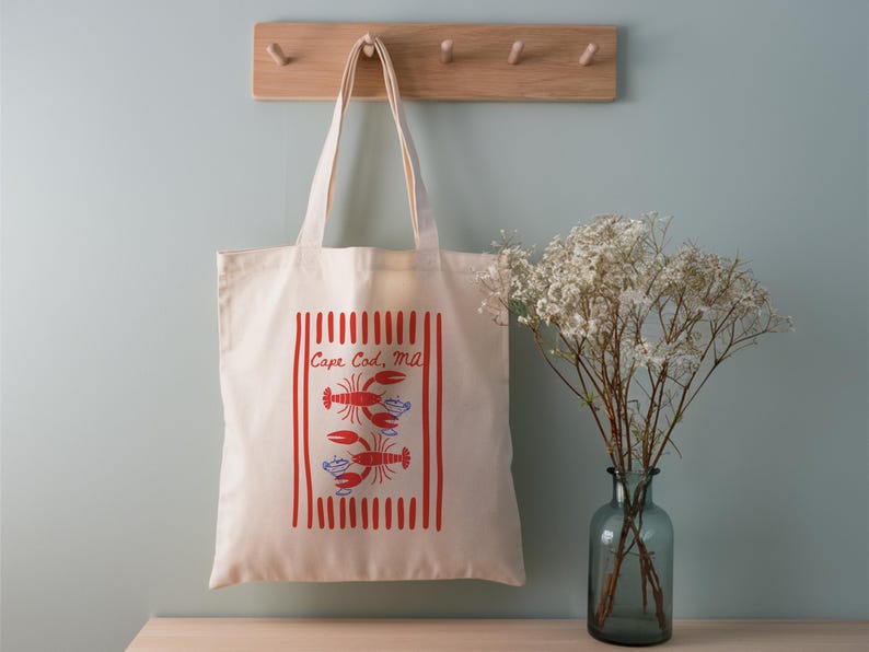 May include: A cream-colored canvas tote bag featuring a red vertical striped rectangular border. Inside the border are two red lobster illustrations and the text Cape Cod, MA in red script. The bag has two long shoulder straps for carrying.