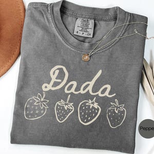 Op de afbeelding: Een grijs t-shirt met het woord "Dada" in een cursief lettertype en vier aardbeienillustraties. Het shirt heeft een ronde hals en korte mouwen. Een zilveren ketting met een hanger ligt op het shirt.