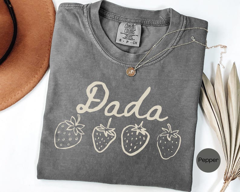 Op de afbeelding: Een grijs t-shirt met het woord "Dada" in een cursief lettertype en vier aardbeienillustraties. Het shirt heeft een ronde hals en korte mouwen. Een zilveren ketting met een hanger ligt op het shirt.