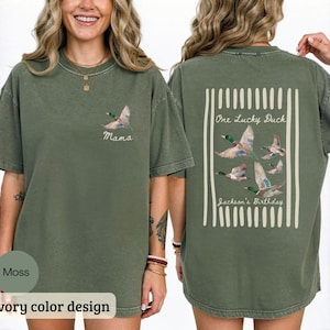 May include: Moss green t-shirt with a duck graphic. The front of the shirt has a duck graphic and the word "Mama". The back of the shirt has a rectangular design with the words "One Lucky Duck" and "Jackson's Birthday" with duck graphics.