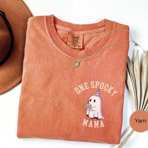 May include: A terracotta-colored short-sleeve t-shirt featuring a graphic of a ghost wearing a party hat and holding a small cake. The text "ONE SPOOKY MAMA" is printed in a curved font around the ghost. The shirt has a crew neckline and a tag.