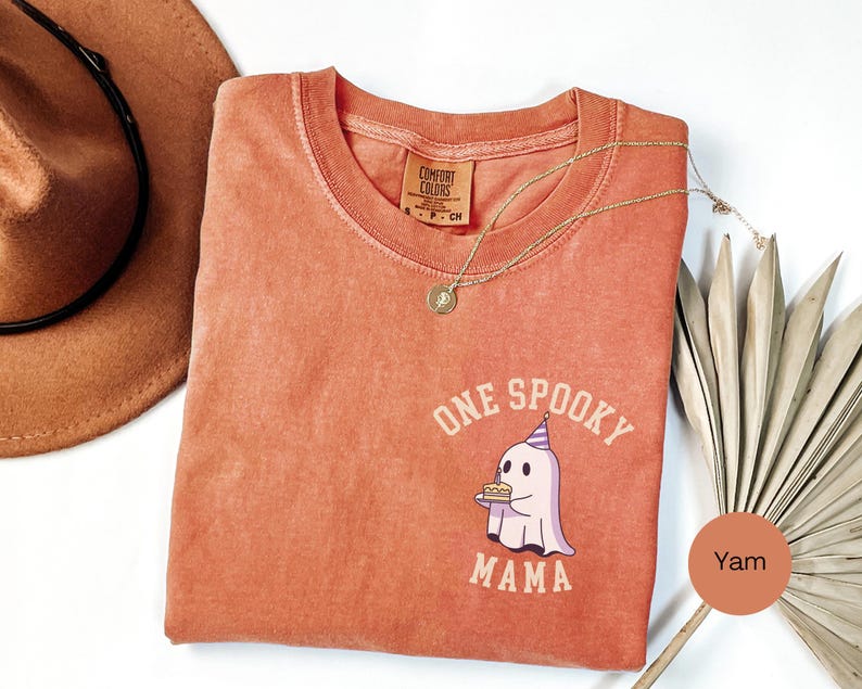 May include: A terracotta-colored short-sleeve t-shirt featuring a graphic of a ghost wearing a party hat and holding a small cake. The text "ONE SPOOKY MAMA" is printed in a curved font around the ghost. The shirt has a crew neckline and a tag.