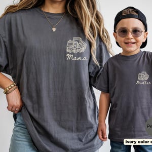 Może przedstawiać: Dwie koszulki w kolorze ciemnoszarym z białą grafiką liniową przedstawiającą wywrotkę. Większa koszulka ma nadruk Mama pod ciężar&oacute;wką, a mniejsza koszulka ma nadruk Brother pod ciężar&oacute;wką wykonany kursywą.