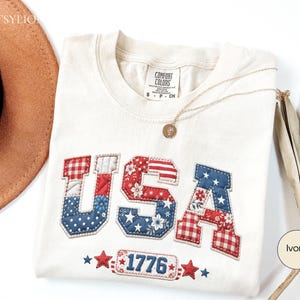 May include: Ivory-colored t-shirt with the word "USA" in red, white, and blue patchwork letters. The number "1776" is below the letters. A gold necklace rests on the shirt. The shirt is a Comfort Colors brand.