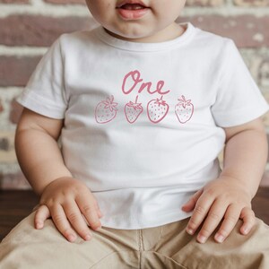 Op de afbeelding: Wit T-shirt met het woord "One" en vier roze aardbeien illustraties. Het shirt wordt door een kind gedragen. Het kind draagt een kaki broek en zit.