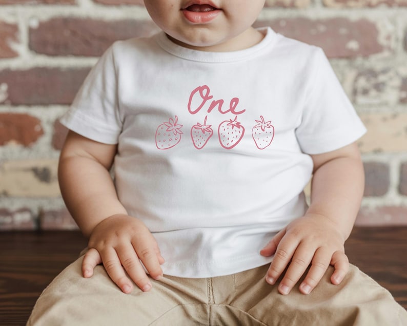 Op de afbeelding: Wit T-shirt met het woord "One" en vier roze aardbeien illustraties. Het shirt wordt door een kind gedragen. Het kind draagt een kaki broek en zit.
